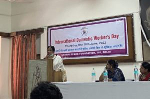 A women standing at a podium speaking. She is wearing a white shawl over her shoulders and a mask sits under her chin. There is another woman sitting at a table next to the podium looking at the woman speaking. There is a big white banner behind the, that says 'International Domestic Workers Day, Thursday 16th June 2022.