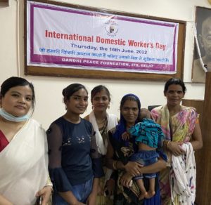 Five women stand in front of a big white banner that says International Domestic Workers Day, Thursday the 16th June 2022. One woman is holding a young child. 