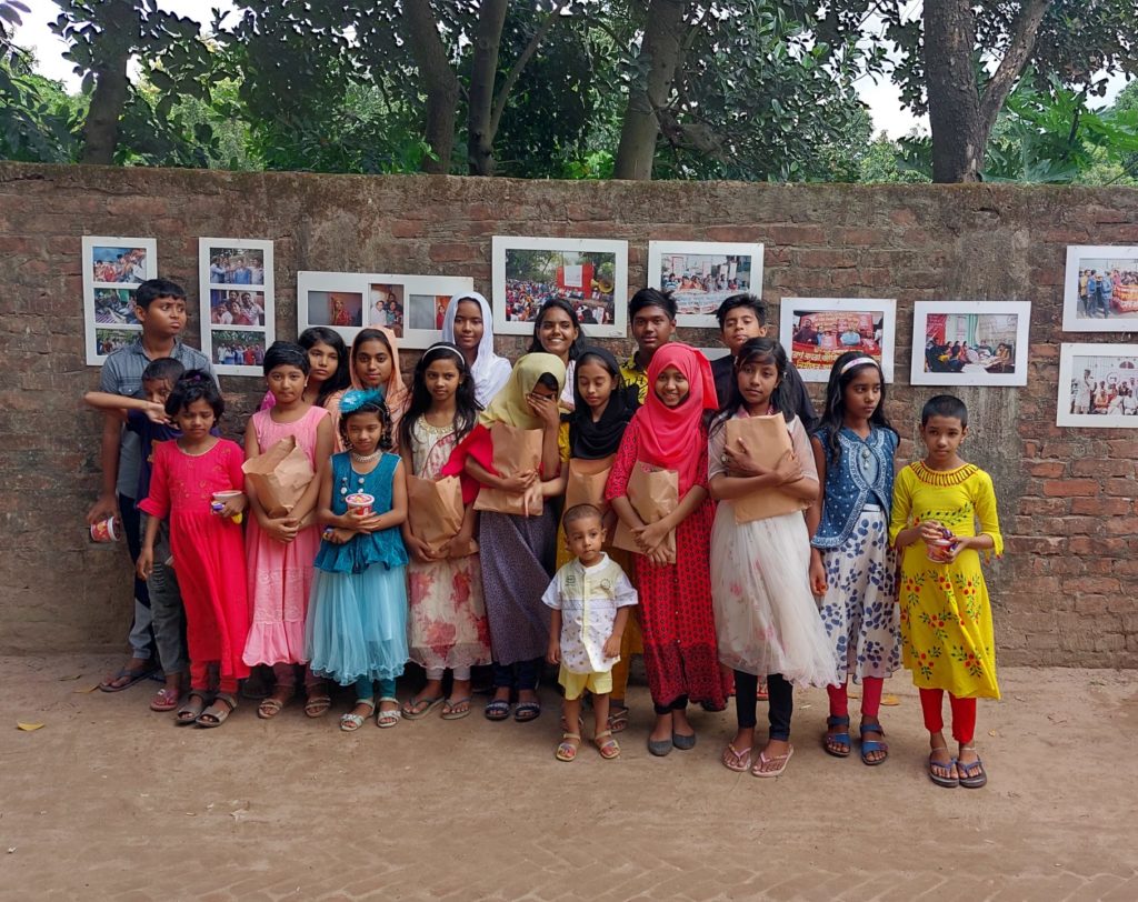 Children of the ready made garment workers stand for a group photo. They are standing in front of a brick wall with photographs on it and trees behind it.