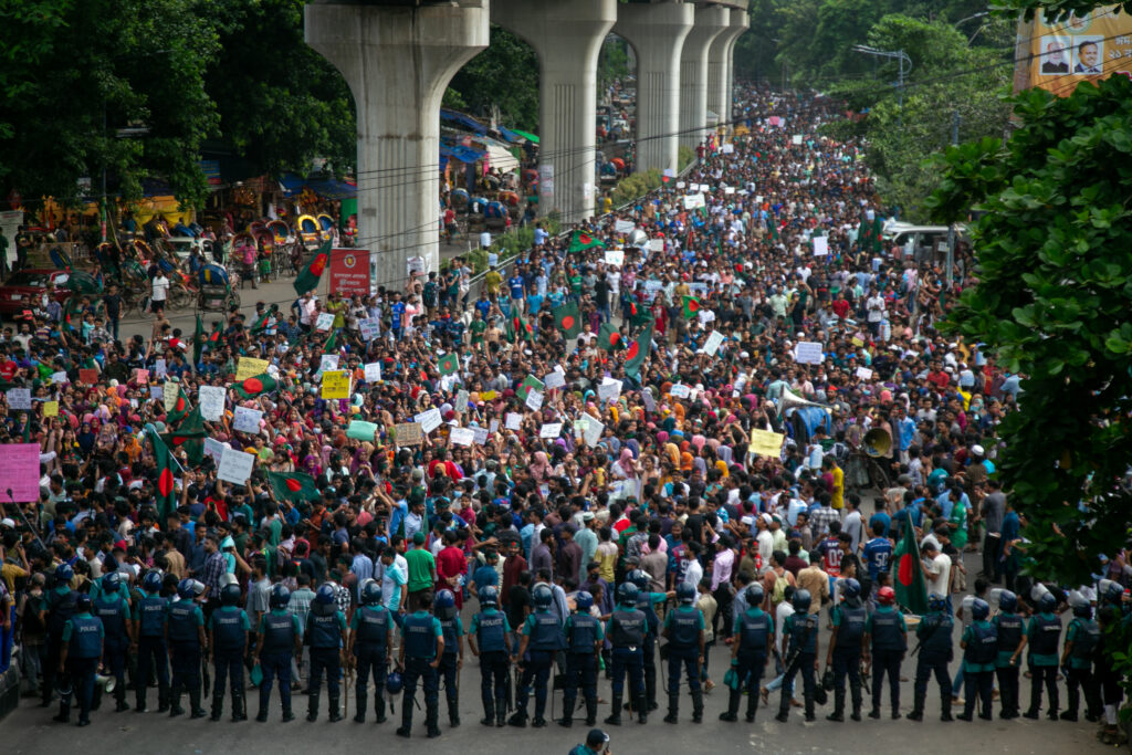 Thousands of students in Dhaka protest on the street, and are blocked by a line of police.